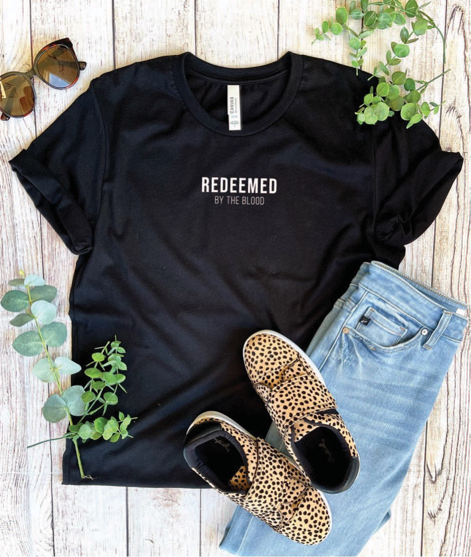 This is a basic back unisex style tee with the words, "REDEEMED BY THE BLOOD," in white. It is a regular fit with a crew neckline. The graphic is featured as a simple crest-size design slightly above the middle of the chest area. 