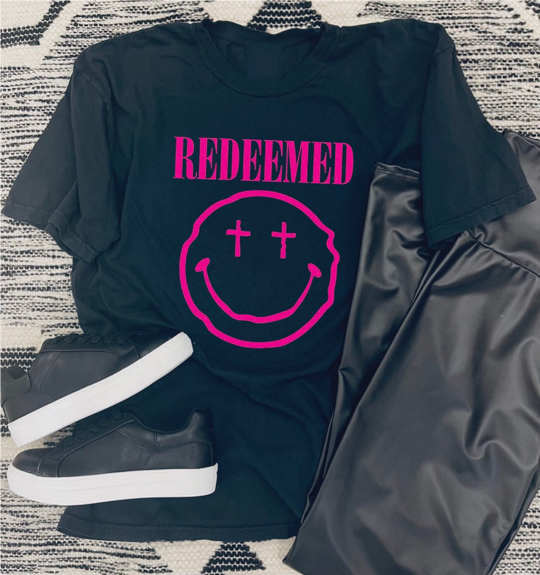 This style is a black unisex tee with a crew neckline. It has a bright neon pink graphic. The graphic features a smiley face with crosses for the eyes. Below, it states REDEEMED in bold lettering. 