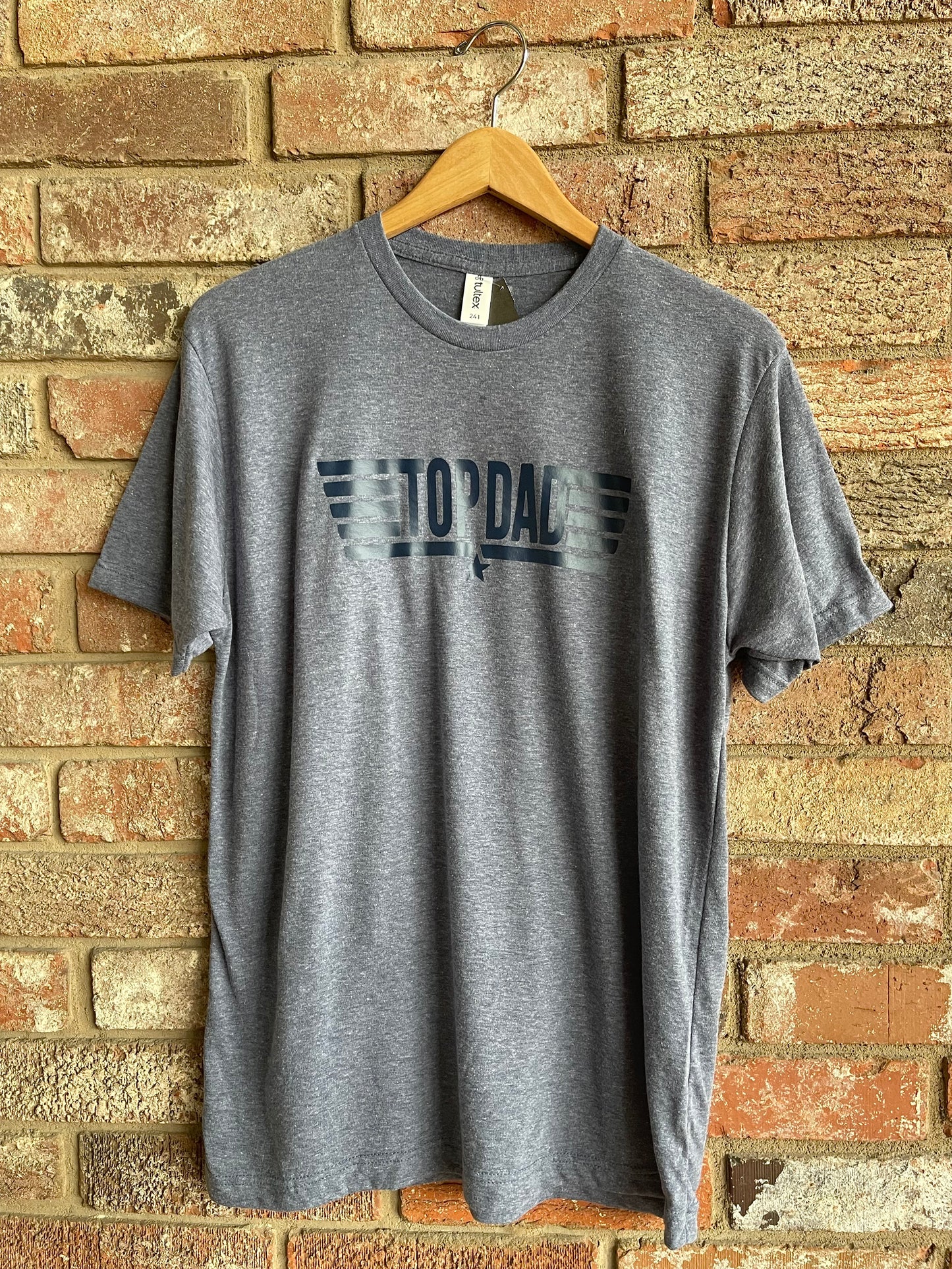 This is a heather blue unisex style tee with a crew neckline. It features a navy blue graphic and lettering, stating, "TOP DAD." It is a regular soft style fit.