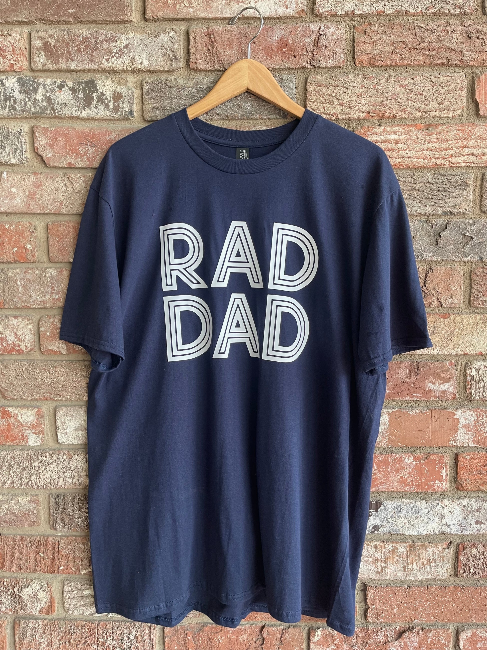 This is a navy blue unisex style tee with lettering, stating, "RAD DAD" on the front. It is a regular fit with a crew neckline. Looking for a fun tee for Father's Day? Look no further!