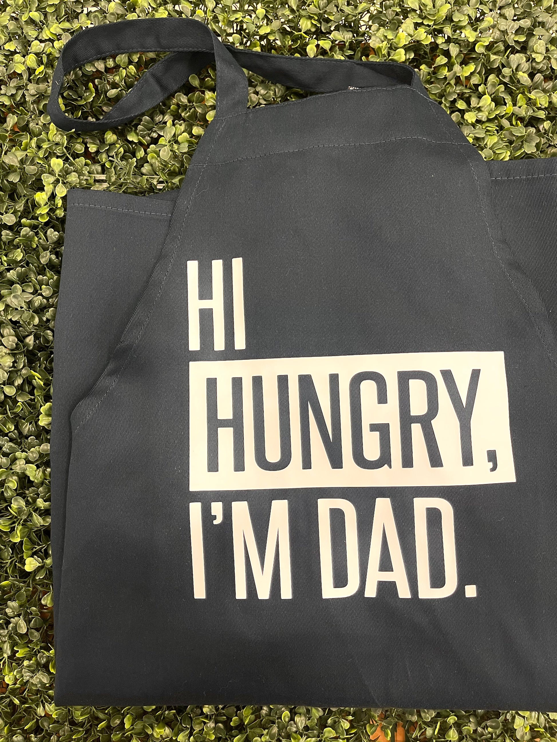 This is a navy blue apron that goes around the neck and has an adjustable tie in the back. It has a white graphic, stating, "HI HUNGRY, I'M DAD." This is a perfect gift option for the dads who love dad jokes!