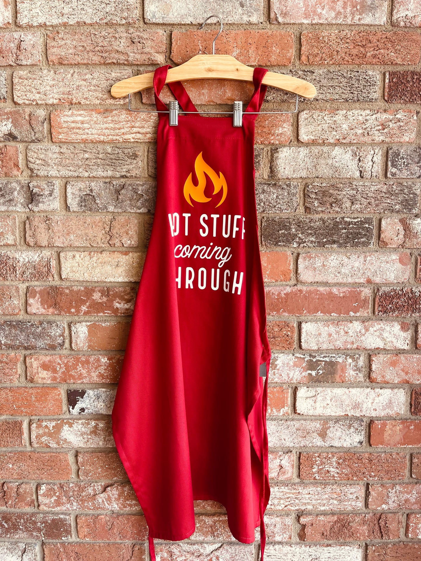 This is a red apron with a functional, adjustable tie that goes around the waist. It has "hot stuff coming through" in white lettering with a yellow flame above the design. 