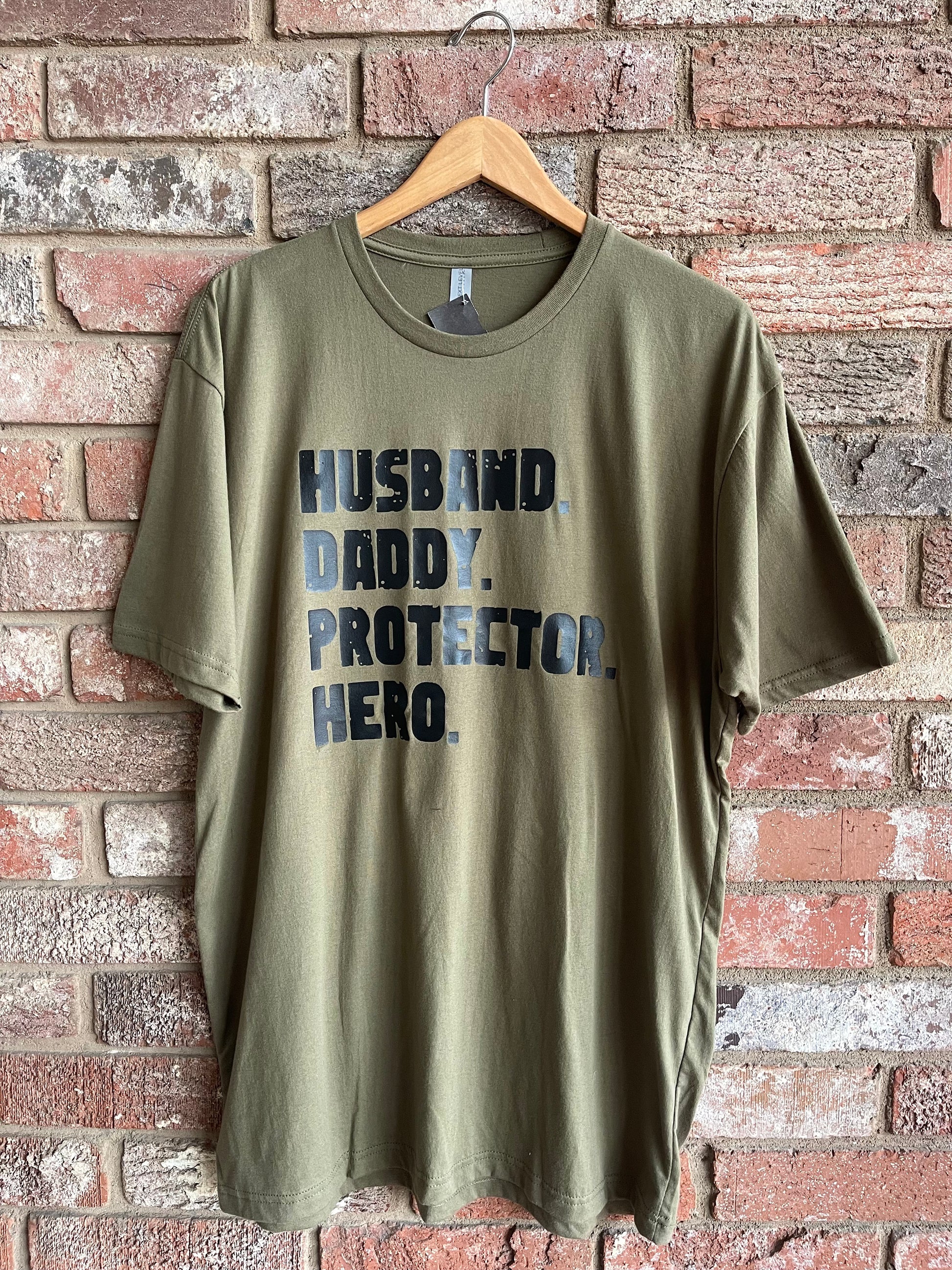This makes an awesome gift option for the hero fathers! This is an olive green soft style tee with black lettering, stating, "HUSBAND, DADDY, PROTECTOR, HERO." This style has a crew neckline and is a regular unisex fit.