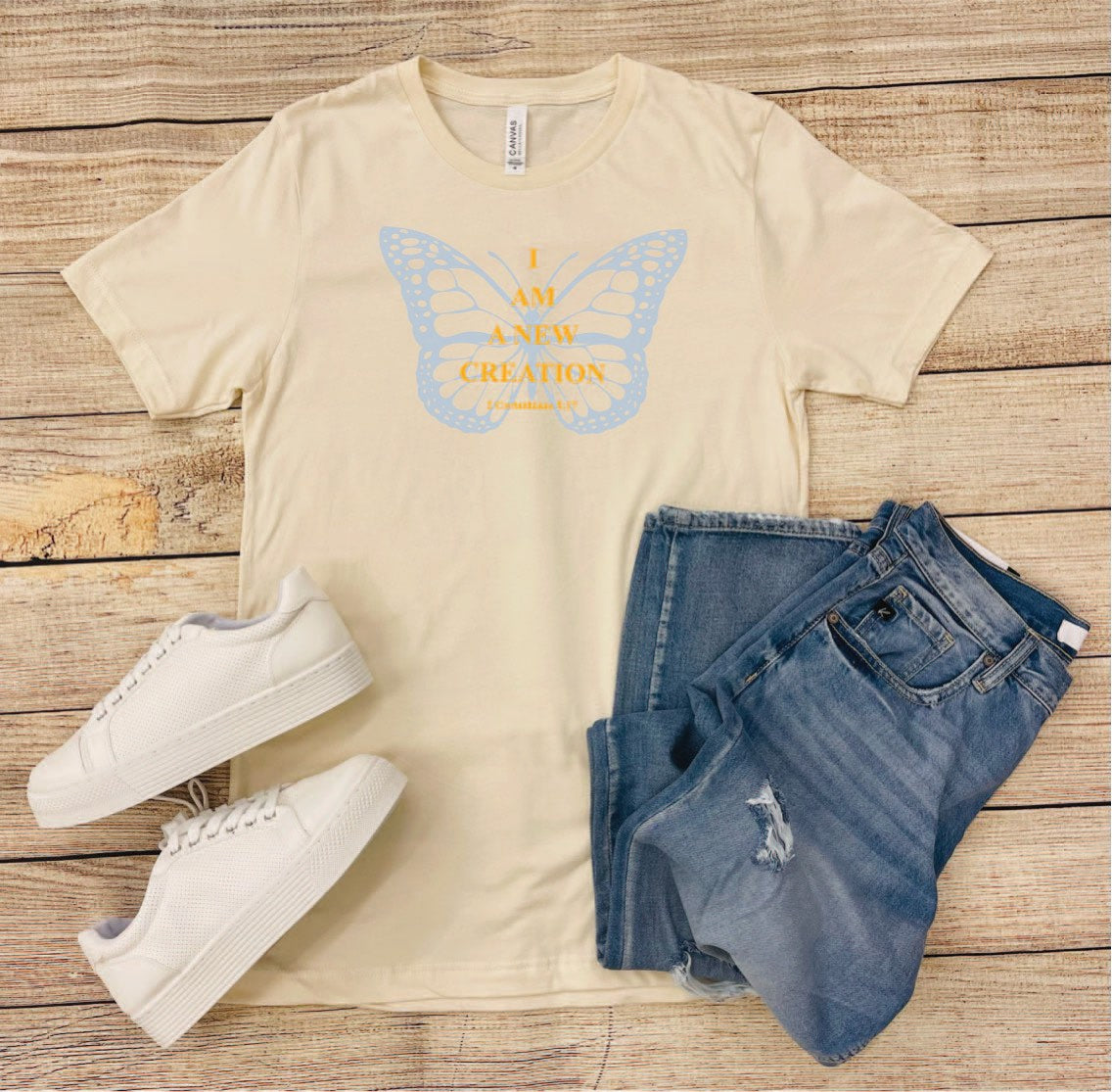 This is a creamy tan tee with a light blue and gold graphic. The graphic features a blue butterfly with the words, "I AM A NEW CREATION," along with the Bible verse underneath. It is a regular unisex style with a crew neckline.